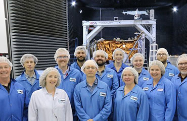 Groupe de 13 personnes en sarraus bleus, souriant, debout, devant l’un des engins spatiaux de la mission de la Constellation RADARSAT dans un entrepôt.