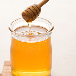 A honey dipper scoops a trail of honey from a filled glass jar.