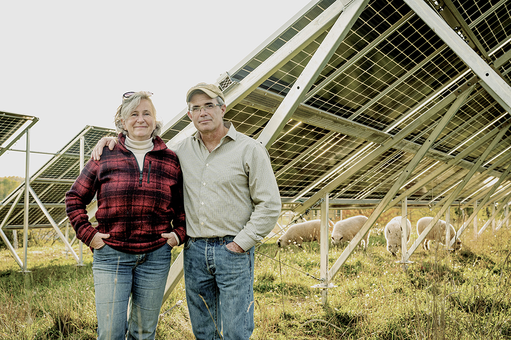 Nancy and her husband standing in front of large solar panel while sheep graze in the background.