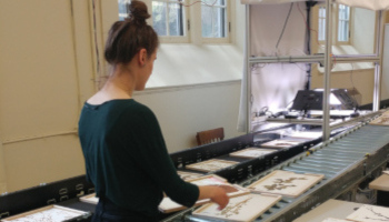 A young woman with her back to the camera places flat, dry-preserved plant specimens from the herbarium onto a conveyor belt that feeds into an automated imaging system.