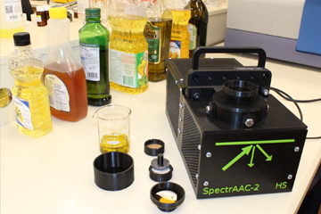 Sur une table, on voit l’appareil SpectrAAC-2, des échantillons d’huile à tester et à l’arrière-plan une rangée de bouteilles d’huiles végétales