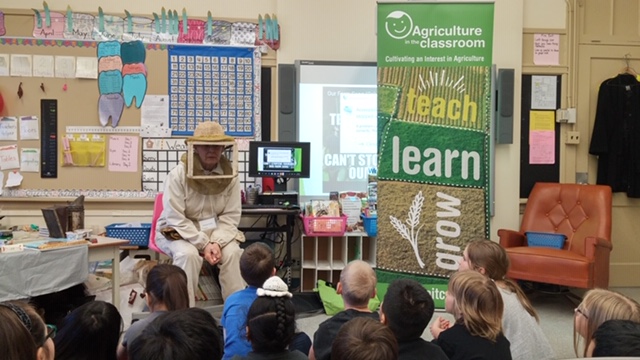 Dressed in a beekeeper suit, speaking with children in a class.
