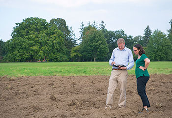 On the right of the photo, Dr. Andrew Davidson and Dr. Catherine Champagne look down at a computer tablet in his hands while standing in a plowed field with trees in the background.
