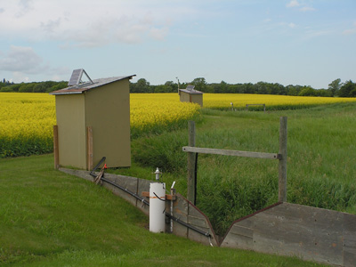 Two monitoring equipment huts and v-notch weirs to channel runoff. There is a field of canola in the background.