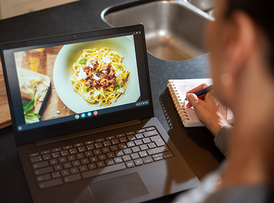 A person writing in a notebook next to a laptop showing a plate full of spaghetti.