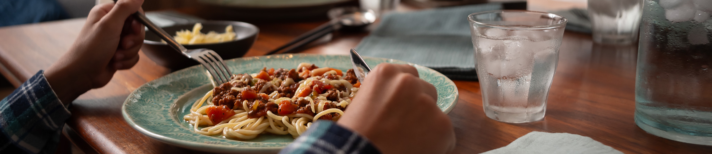 Hands holding a fork and knife over a plate of spaghetti at a dinner table.