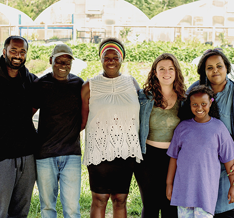 Jacqueline and Noel with a man, two women, and a young girl smiling at the camera with a field of vegetables and greenhouses in the background