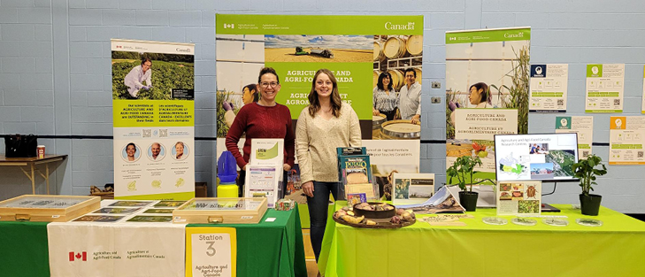 Two women standing next to each other behind tables covered in potatoes, bee pictures and bee displays.