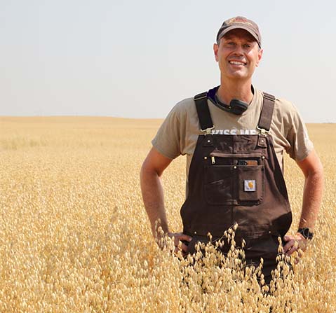 A man in overalls standing in a field smiling to the camera.