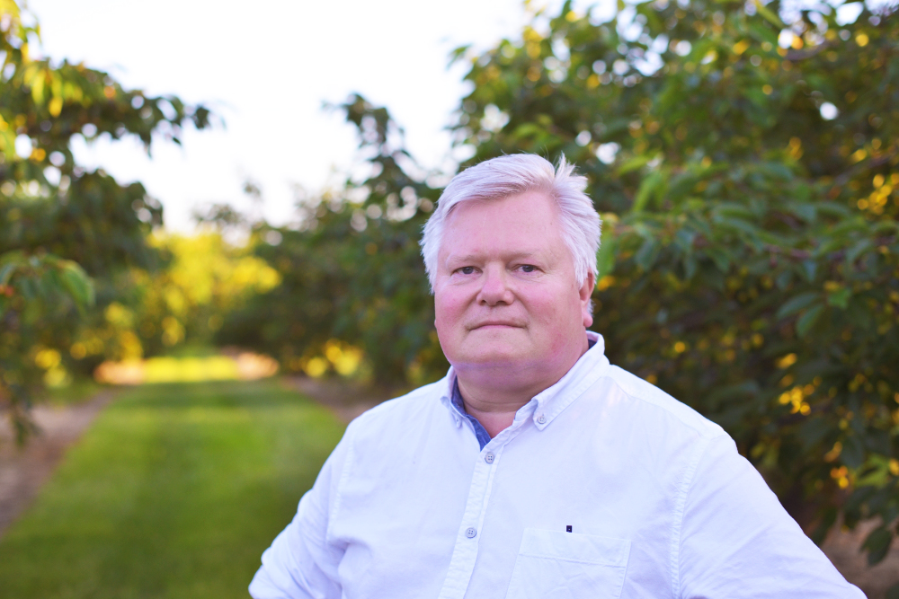 Paul Moyer in a white shirt standing next to trees, looking towards the camera.