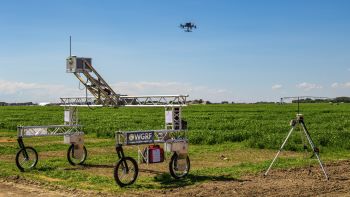 A drone in the bright blue sky, over green field plots, with a tall cart-like structure in the foreground